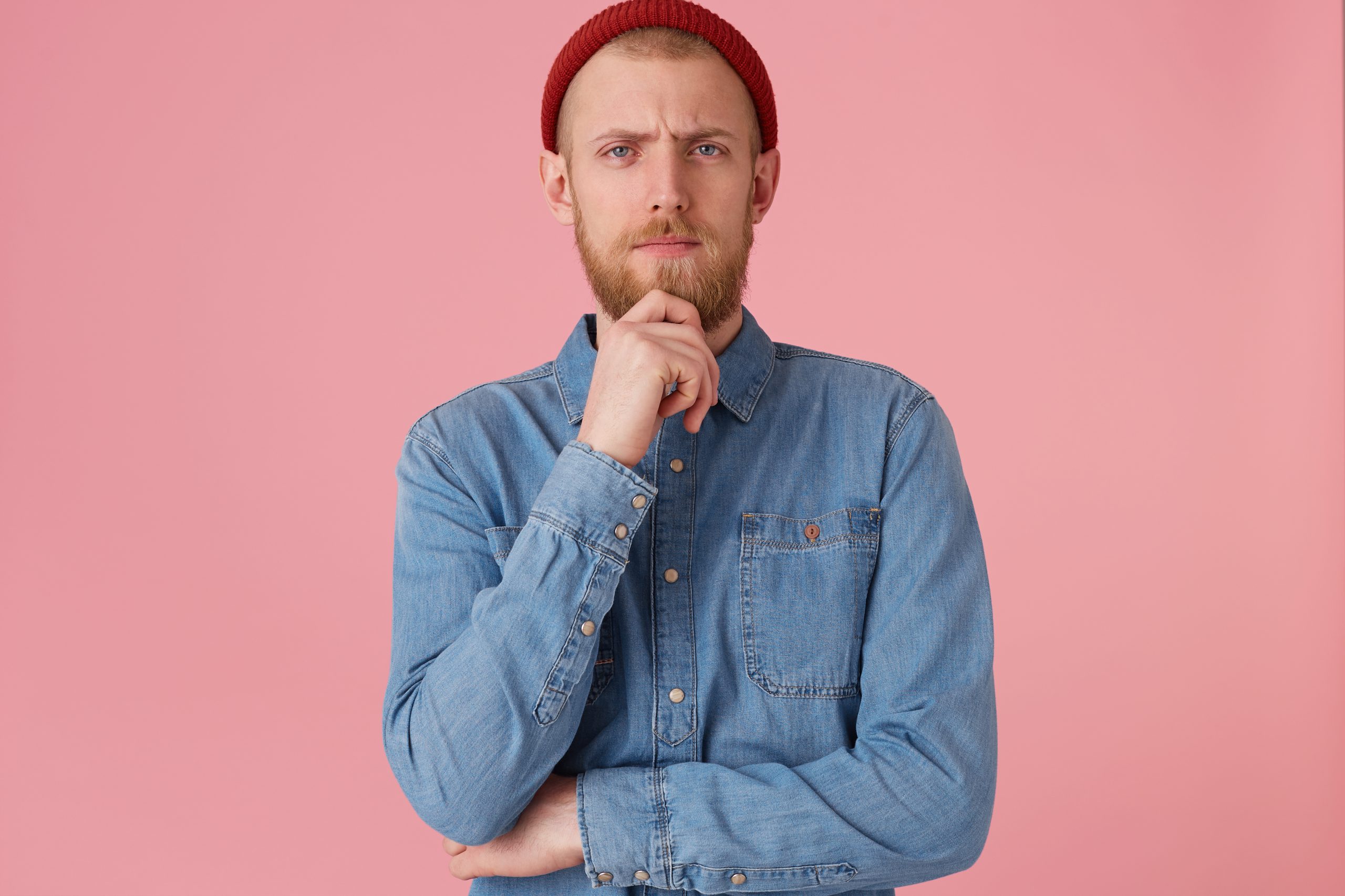 A handsome thoughtful bearded guy looking camera holding his chin, thinks about his future, make plans, dreams, feels confident in his abilities, isolated over pink background